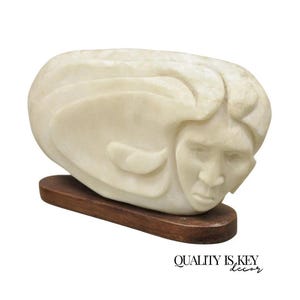 May include: A carved stone sculpture of a human face and hair, in a light cream color, mounted on a dark brown wooden base. The sculpture has a smooth, polished finish. The text "QUALITY IS KEY decor" is at the bottom.