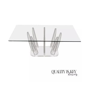 May include: A clear glass top dining table with a unique acrylic base featuring a geometric design. The base is made of multiple acrylic pieces that are arranged in a symmetrical pattern.