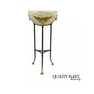 May include: A decorative stand with a round, white, mosaic bowl with gold accents. The stand is made of black metal with gold feet.