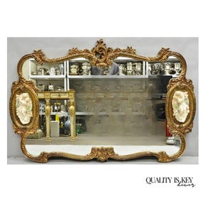 May include: Ornate gold-framed mirror with a rectangular shape. The frame features intricate carvings and two oval panels with white relief figures. The mirror reflects a room with shelves and decorative items. The text "QUALITY IS KEY decor" is at the bottom.