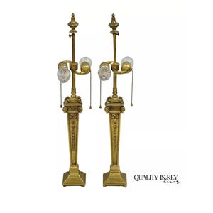 May include: Pair of antique gold-toned table lamps. Each lamp features a detailed base, a central column with ornate carvings, and a three-light fixture. The lamps have pull chains and decorative finials. The text "QUALITY IS KEY decor" is visible.