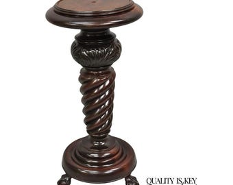 Puede incluir: Un soporte para plantas de estilo antiguo, de color marrón oscuro, con una parte superior e inferior redondas. El soporte presenta un diseño floral tallado y una columna central retorcida en espiral. La base tiene tres patas de garra.