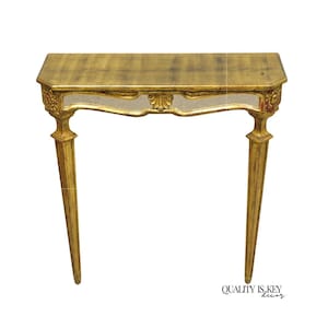May include: A gold-colored console table with a rectangular top and two slender, tapered legs. The table features ornate detailing, including a decorative floral motif and a mirrored section below the tabletop. The words "QUALITY IS KEY DECOR" are printed on the bottom right.
