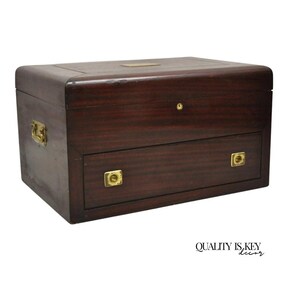 May include: A dark mahogany wooden box with a drawer, brass hardware, and a small brass plate on the top. The box has a rectangular shape with rounded edges and a keyhole on the front. The text "QUALITY IS KEY decor" is at the bottom.