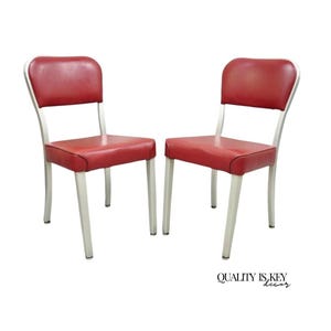 May include: Two vintage chairs with red seats and backs, and silver-colored metal frames. The chairs have a classic design, suitable for a kitchen or dining room. The chairs are shown against a white background. The text "QUALITY IS KEY decor" is visible.