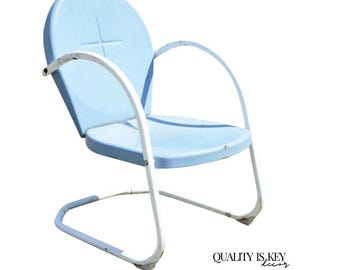 May include: A vintage light blue metal chair with white armrests. The chair has a curved back with a cross design and a curved base. The chair is a retro style, perfect for a patio or garden.