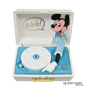 May include: Vintage General Electric record player in a white case with a blue interior. The lid features a Mickey Mouse illustration and the text "Hi kids!". The record player has a white turntable and a white hand pointing towards the record.