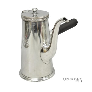 May include: A silver-colored coffee pot with a black handle. The pot has a lid with a small knob and a spout. A decorative emblem is visible on the side. The pot is made of metal and has a shiny finish.