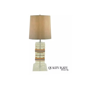 May include: A clear acrylic table lamp with a stacked base and a beige fabric lampshade. The base has brown and black accents.