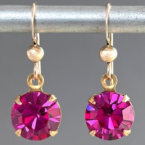 May include: A pair of gold-colored drop earrings featuring large, round, fuchsia-colored crystals. The crystals are faceted, catching the light. The earrings hang from a simple gold hook and a small gold bead.