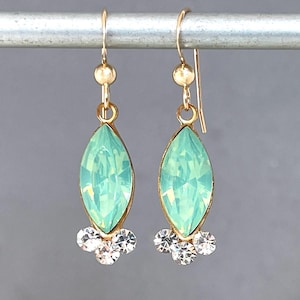 May include: A pair of gold-toned drop earrings featuring a central marquise-cut mint green crystal. Each earring is accented with three small, clear round crystals at the base and hangs from a gold-toned hook with a small gold bead detail.