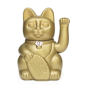 May include: A gold-colored Maneki-Neko, or lucky cat, figurine with its right paw raised. The cat is holding a gold coin with Japanese characters. The cat has a white flower on its collar.