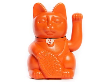 以下が含まれることがあります： オレンジ色のセラミック製の招き猫の置物。片方の足が上がり、もう片方の足には魚を持っています。首には白い花のチャームが付いています。