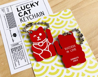 May include: Red lucky cat keychain with white details. The keychain features the text "Success in Love" and "Diminuto Cielo".