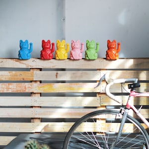 May include: A row of six colorful lucky cat figurines, also known as Maneki-Neko, are displayed on a wooden pallet wall. The cats are in various colors, including blue, red, yellow, pink, green, and orange. A bicycle is partially visible in the background.