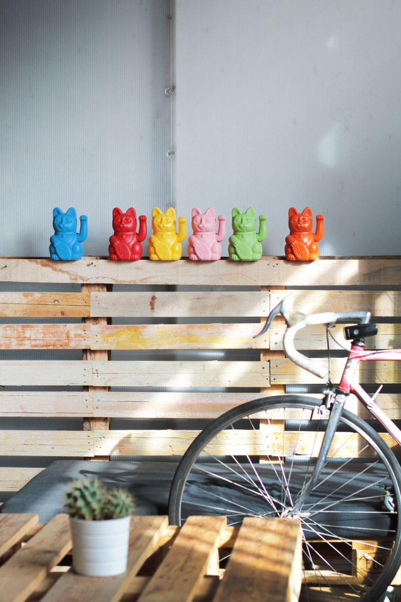 May include: A row of six colorful lucky cat figurines, also known as Maneki-Neko, are displayed on a wooden pallet wall. The cats are in various colors, including blue, red, yellow, pink, green, and orange. A bicycle is partially visible in the background.