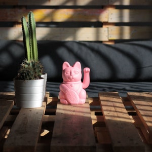 May include: A pink lucky cat figurine with its right paw raised, sitting on a wooden pallet with a cactus plant in a white pot to the left.