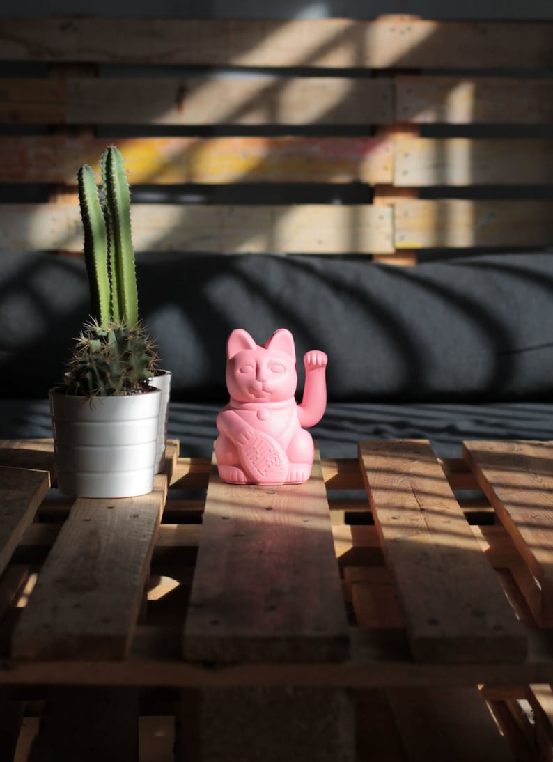May include: A pink lucky cat figurine with its right paw raised, sitting on a wooden pallet with a cactus plant in a white pot to the left.
