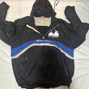 May include: Black Champion windbreaker jacket with a hood and a zipper. The jacket has a blue and white stripe across the chest and the text "BATTLEHAWKS" and "Champion" printed on it. The jacket has a drawstring at the bottom and a small pocket.