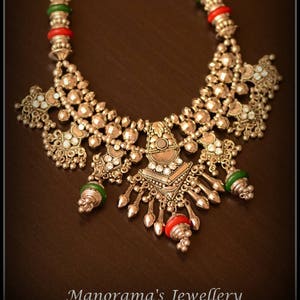 May include: A silver necklace with a large, ornate pendant featuring a geometric design and small, clear stones. The necklace is accented with green and red beads.