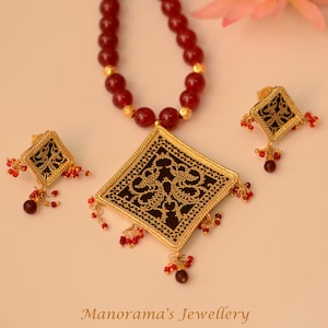 May include: A gold necklace with a square pendant featuring a black filigree design. The necklace is made of red beads and has matching earrings with a similar design.