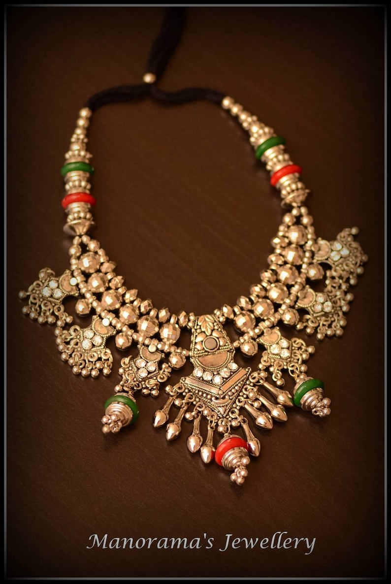 May include: A silver necklace with a large, ornate pendant featuring a geometric design and small, clear stones. The necklace is accented with green and red beads.