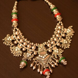 May include: A silver necklace with a large, ornate pendant featuring a mirror-like center and intricate details. The necklace is adorned with green and red beads and has a black cord for closure. "Manorama's Jewellery" is written in the bottom of the image.