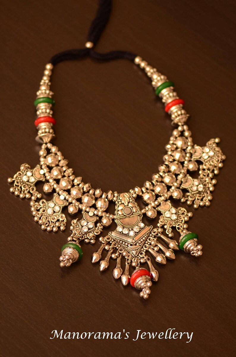May include: A silver necklace with a large, ornate pendant featuring a mirror-like center and intricate details. The necklace is adorned with green and red beads and has a black cord for closure. "Manorama's Jewellery" is written in the bottom of the image.