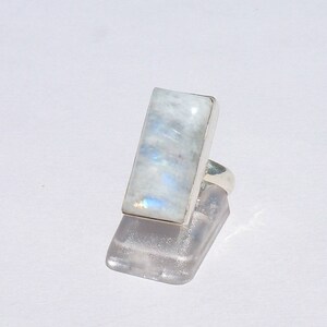 May include: A rectangular moonstone ring with a silver band. The moonstone is a light, opalescent color with blue and white hues. The ring is set on a clear display stand.