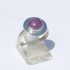 Può includere: Un anello d'argento con una pietra preziosa rotonda di colore viola incastonata in una montatura d'argento. L'anello è su un espositore trasparente.