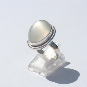 Op de afbeelding: Een zilveren ring met een grote, ovale, lichtgrijze edelsteen. De edelsteen is gezet in een zilveren bezel met een dubbel bandontwerp. De ring wordt gepresenteerd op een transparante plastic standaard tegen een witte achtergrond.