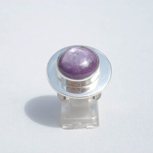 Puede incluir: Un anillo de plata con una gran piedra preciosa redonda y pulida de color púrpura. La piedra preciosa está engastada en un bisel y el anillo tiene una banda ancha y plana. El anillo se exhibe sobre un soporte transparente sobre un fondo blanco.