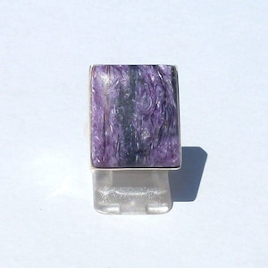 Könnte beinhalten: Ein rechteckiger Ring mit einem großen, polierten, lila Edelstein mit dunklen Streifen, gefasst in einem silberfarbenen Metallband. Der Ring wird auf einem klaren, rechteckigen Ständer vor weißem Hintergrund präsentiert.