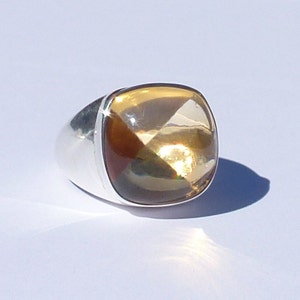 Può includere: Un anello d'argento con una grande pietra preziosa color ambra, tagliata a forma quadrata. La gemma ha molteplici sfaccettature che riflettono la luce, creando un effetto scintillante. La fascia dell'anello è liscia e lucida.