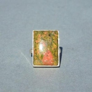 Puede incluir: Un anillo rectangular con una piedra natural en tonos verdes, naranjas y amarillos. La piedra está engastada en una banda de metal plateado. El anillo tiene un diseño sencillo y elegante.