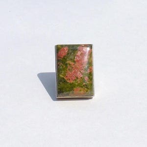 Peut inclure: Une pierre précieuse d'unakite rectangulaire avec une bordure en métal argenté. La pierre présente un mélange de couleurs vertes et roses. L'unakite est posée sur un fond blanc.