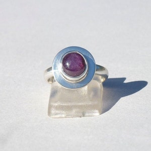 Może przedstawiać: Srebrny pierścionek z fioletowym kamieniem szlachetnym osadzonym w oprawie typu bezel. Pierścionek znajduje się na białym stojaku.