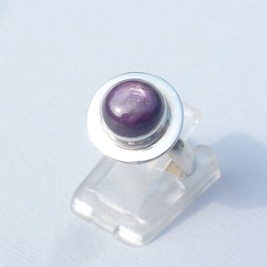 Puede incluir: Un anillo de plata con una gema grande, redonda y de color púrpura intenso. La gema está engastada en un bisel circular. El anillo se exhibe sobre un soporte transparente. La banda del anillo es fina y sencilla.