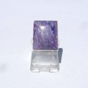 May include: A rectangular ring featuring a large, polished purple gemstone with white and gray veining. The stone is set in a silver band. The ring is displayed on a clear plastic stand against a white background.