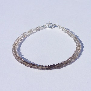 May include: A beaded bracelet featuring small, faceted smoky quartz stones. The beads transition from light to dark brown, strung together with a silver clasp. This piece is a delicate accessory.