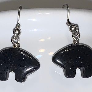 Peut inclure: Une paire de boucles d'oreilles pendantes en argent avec des pierres en forme d'ours noir. Les pierres ont une texture scintillante et brillante.