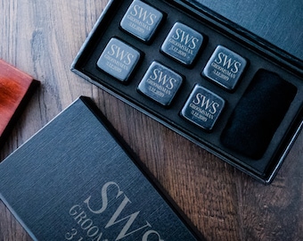 May include: A black gift box with a black velvet interior containing six black square whiskey stones. The stones are engraved with the initials "SWS" and the word "Groomsman". The box also includes a black velvet pouch. The box is open and the stones are visible. The box is sitting on a wooden surface.
