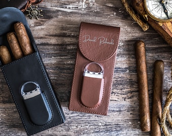 May include: An overhead shot features a black leather cigar case with three cigars, a brown leather case with a lighter, and two loose cigars. The brown case is embossed with "David Richards". A compass and rope are also visible.