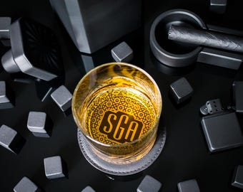 May include: A glass of amber-colored whiskey with ice cubes in a rocks glass. The glass has a gold-colored coaster with the initials "SGA" printed on it. The glass is surrounded by black square ice cubes on a black surface.