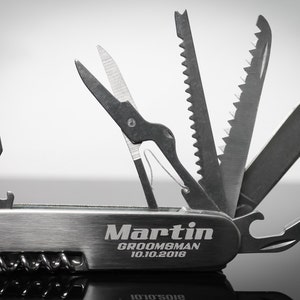 May include: A silver multi-tool with various implements extended, including scissors, a saw, and a knife. The tool is engraved with the name "Martin", the word "Groomsman", and the date "10.10.2018".