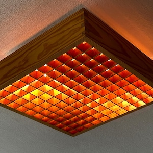 Puede incluir: Luminaria de techo cuadrada de madera con una rejilla de aberturas en forma de diamante. La luminaria es de madera clara y emite un brillo cálido y anaranjado. El diseño es geométrico y moderno, adecuado para la iluminación interior.