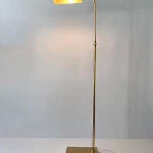 Puede incluir: Una l&aacute;mpara de pie de lat&oacute;n con una base rectangular y un poste vertical y delgado. La l&aacute;mpara tiene un brazo curvo que sostiene una l&aacute;mpara rectangular. La luz est&aacute; encendida, emitiendo un brillo c&aacute;lido. La l&aacute;mpara mide unos 152 cm de alto.