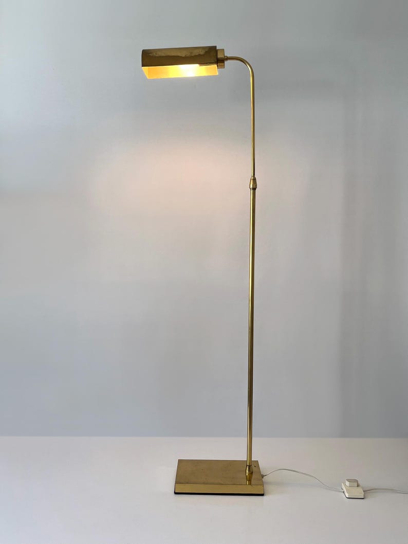 Puede incluir: Una l&aacute;mpara de pie de lat&oacute;n con una base rectangular y un poste vertical y delgado. La l&aacute;mpara tiene un brazo curvo que sostiene una l&aacute;mpara rectangular. La luz est&aacute; encendida, emitiendo un brillo c&aacute;lido. La l&aacute;mpara mide unos 152 cm de alto.