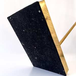 Puede incluir: Un soporte de metal dorado con una base rectangular cubierta de fieltro negro. El soporte tiene una fina varilla de metal dorado que se extiende hacia arriba desde la base.