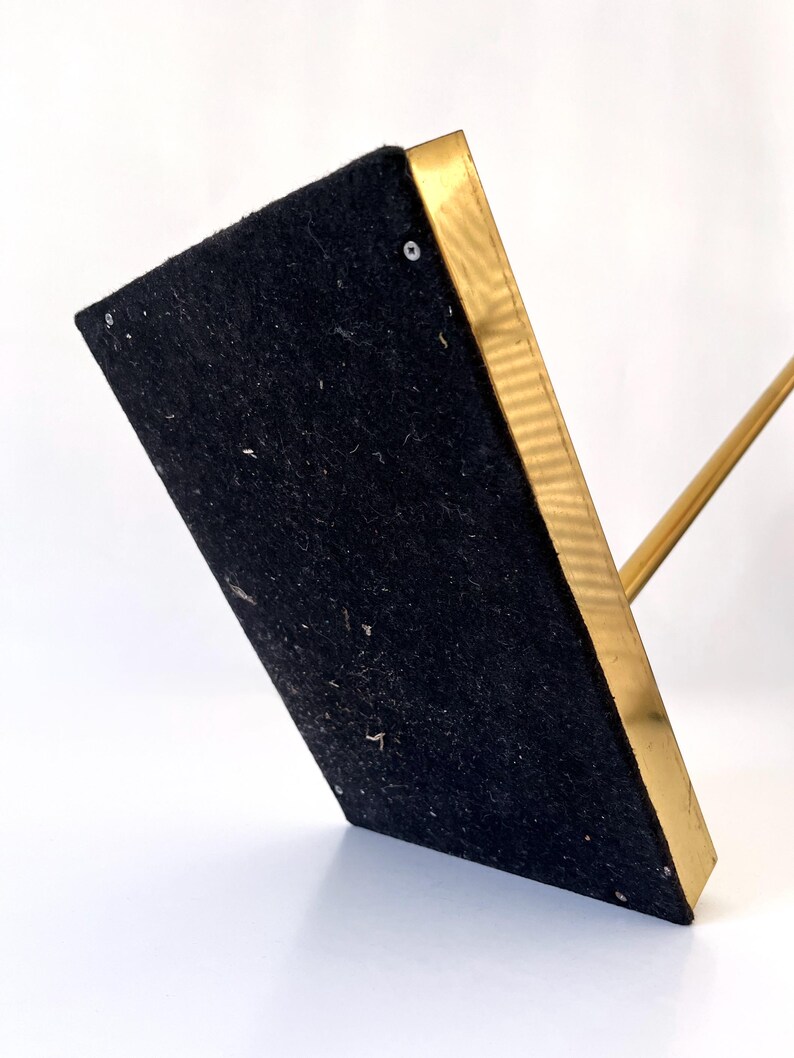 Puede incluir: Un soporte de metal dorado con una base rectangular cubierta de fieltro negro. El soporte tiene una fina varilla de metal dorado que se extiende hacia arriba desde la base.